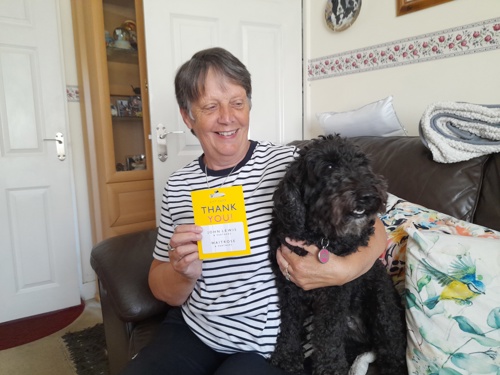 A photo of Mary, prize winner for the Home Safety survey, with her dog. She is holding a gift card and cuddling her dog at the same time. They are sitting on the sofa.
